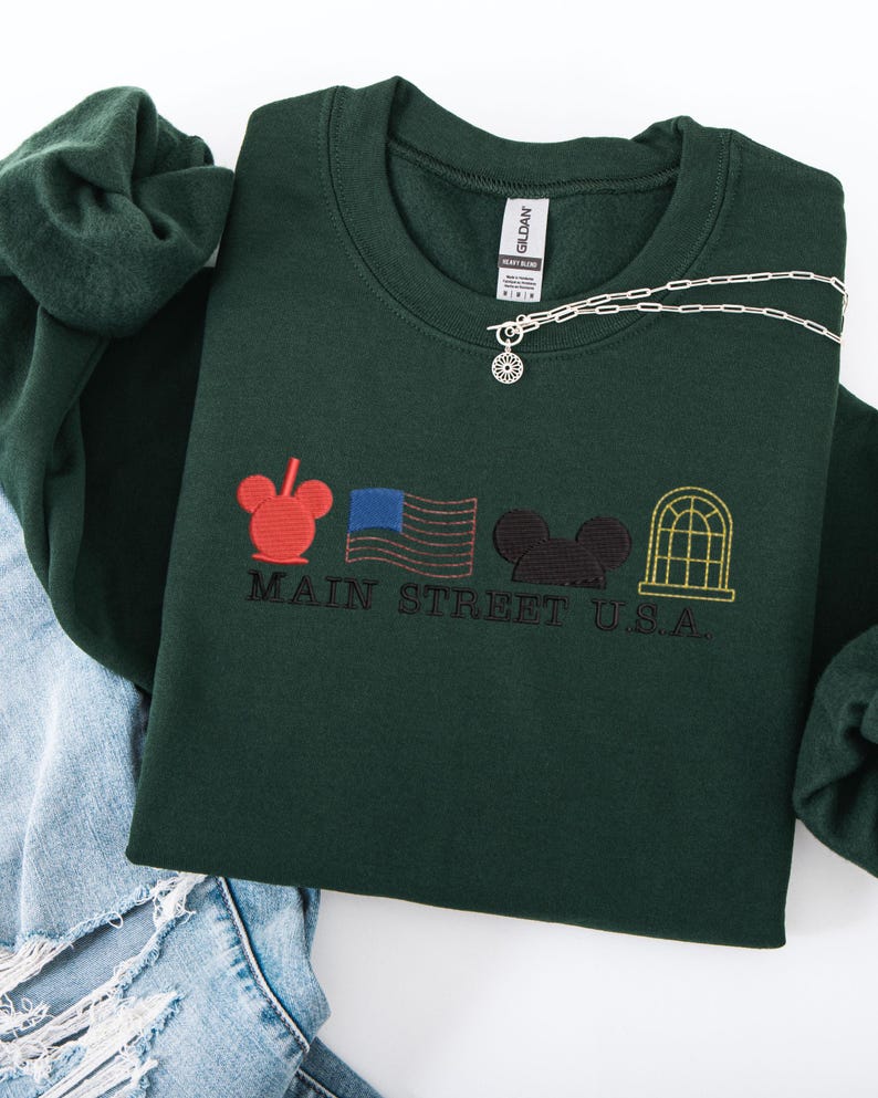 K&ouml;nnte beinhalten: Dunkelgr&uuml;nes Sweatshirt mit gesticktem Main Street U.S.A. Design. Es zeigt einen roten Mickey Mouse-Kopf, eine blaue amerikanische Flagge, einen schwarzen Mickey Mouse-Kopf und ein gelbes Fenster. Eine silberne Halskette mit Sonnenanh&auml;nger liegt darauf.