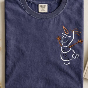 May include: A navy blue Comfort Colors t-shirt with a line drawing of Olaf from Frozen. The design features white, brown, and orange details. The shirt has a crew neck and a small tag on the collar.