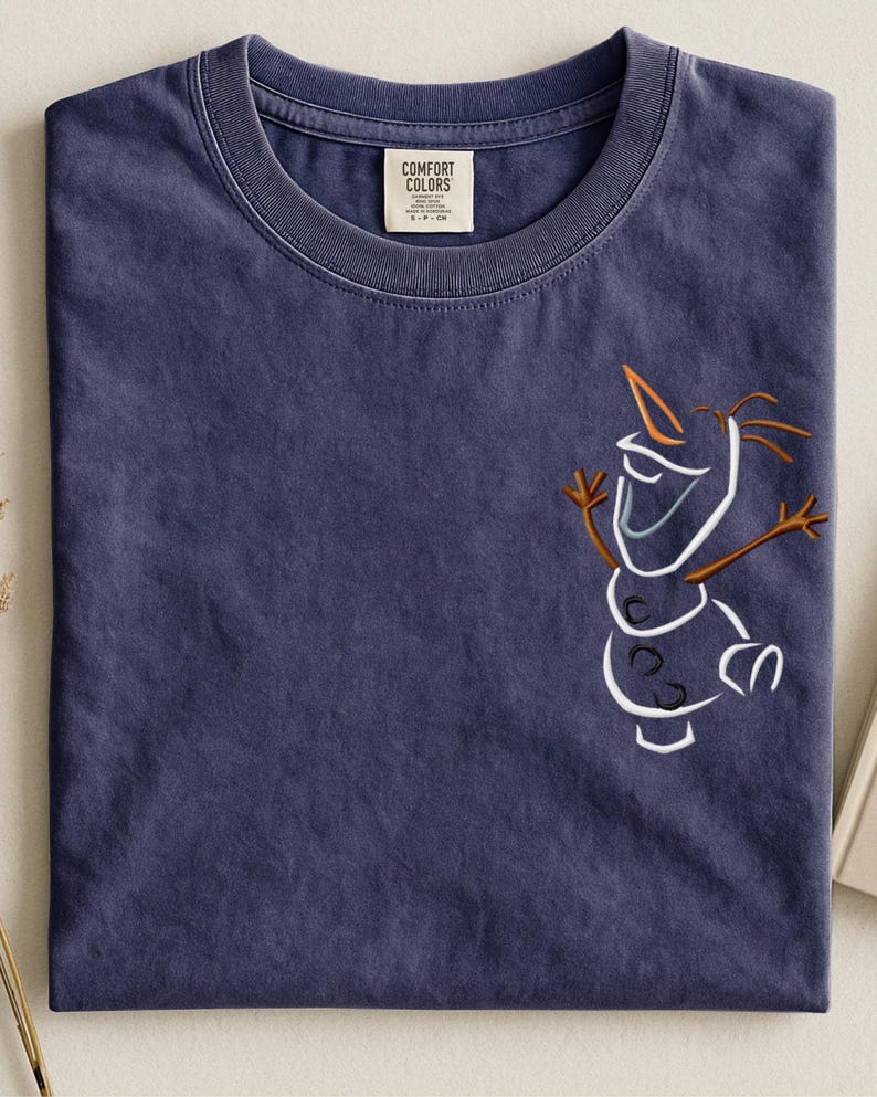 May include: A navy blue Comfort Colors t-shirt with a line drawing of Olaf from Frozen. The design features white, brown, and orange details. The shirt has a crew neck and a small tag on the collar.