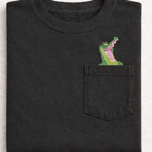 May include: Black t-shirt with a pocket featuring an embroidered cartoon alligator with an open mouth. The alligator is green with a pink mouth and tongue. The shirt is folded neatly.