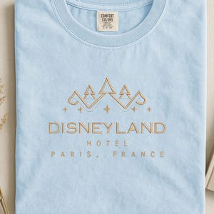 May include: Light blue t-shirt with the text "DISNEYLAND HOTEL PARIS, FRANCE" in gold lettering. The shirt features a gold crown design above the text. The shirt is made by Comfort Colors.