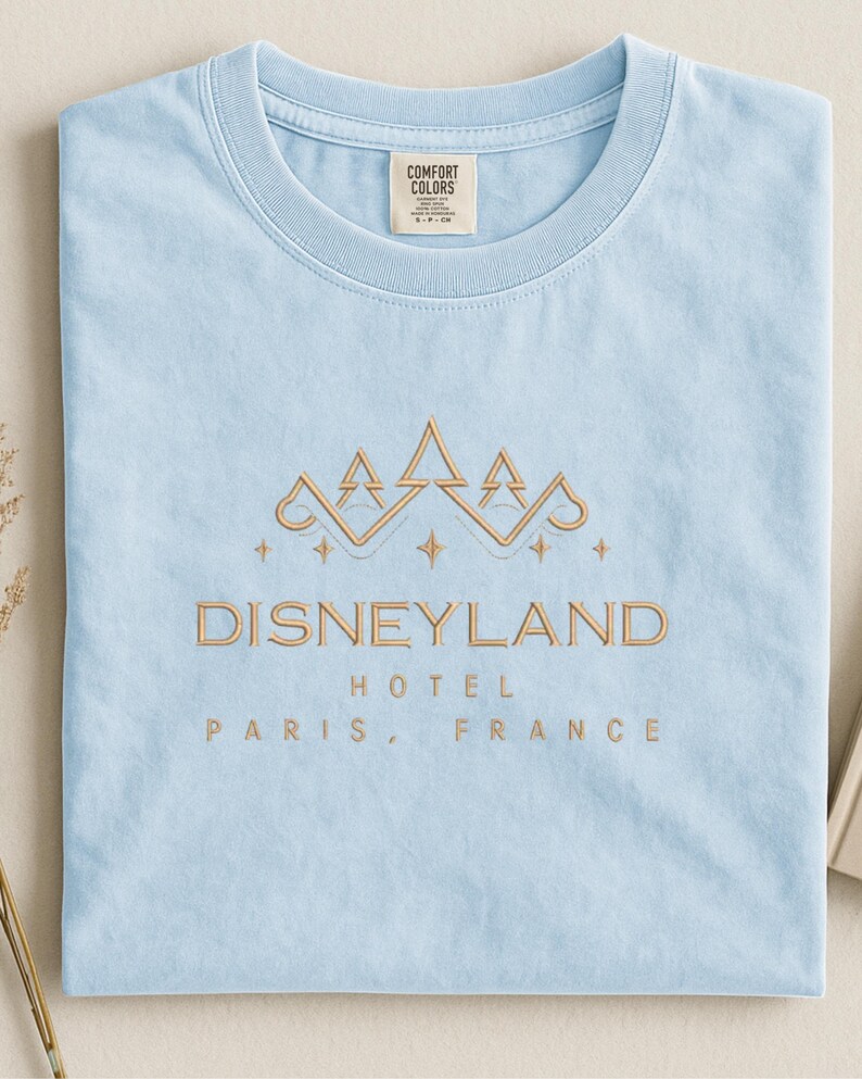 May include: Light blue t-shirt with the text "DISNEYLAND HOTEL PARIS, FRANCE" in gold lettering. The shirt features a gold crown design above the text. The shirt is made by Comfort Colors.