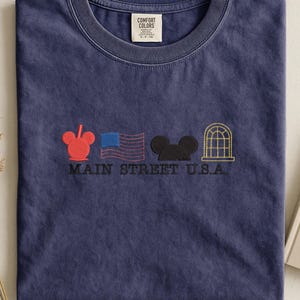 K&ouml;nnte beinhalten: Ein marineblaues T-Shirt mit Rundhalsausschnitt. Das Shirt zeigt gestickte Designs eines roten Mickey Mouse-Kopfes, einer US-Flagge, eines schwarzen Mickey Mouse-Kopfes und eines goldenen Fensters. Die Worte "MAIN STREET U.S.A." sind darunter gestickt.