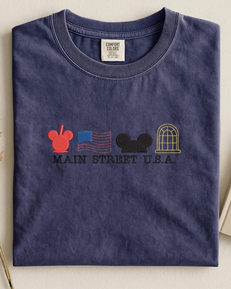 K&ouml;nnte beinhalten: Ein marineblaues T-Shirt mit Rundhalsausschnitt. Das Shirt zeigt gestickte Designs eines roten Mickey Mouse-Kopfes, einer US-Flagge, eines schwarzen Mickey Mouse-Kopfes und eines goldenen Fensters. Die Worte "MAIN STREET U.S.A." sind darunter gestickt.