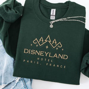 May include: A dark green sweatshirt with the text "DISNEYLAND HOTEL PARIS, FRANCE" in gold lettering. A silver necklace with a sun pendant rests on the sweatshirt. The sweatshirt is folded on top of a pair of blue jeans.