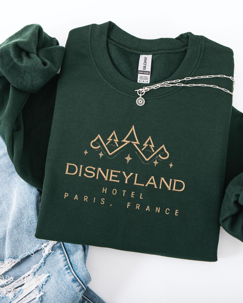 May include: A dark green sweatshirt with the text "DISNEYLAND HOTEL PARIS, FRANCE" in gold lettering. A silver necklace with a sun pendant rests on the sweatshirt. The sweatshirt is folded on top of a pair of blue jeans.