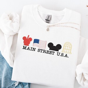 K&ouml;nnte beinhalten: Wei&szlig;es Sweatshirt mit gesticktem Main Street U.S.A. Design. Es zeigt einen roten Mickey Mouse Kopf, eine amerikanische Flagge, schwarze Mickey Mouse Ohren und ein goldenes Fenster. Eine silberne Halskette mit einem kleinen Anh&auml;nger liegt auf dem Shirt.