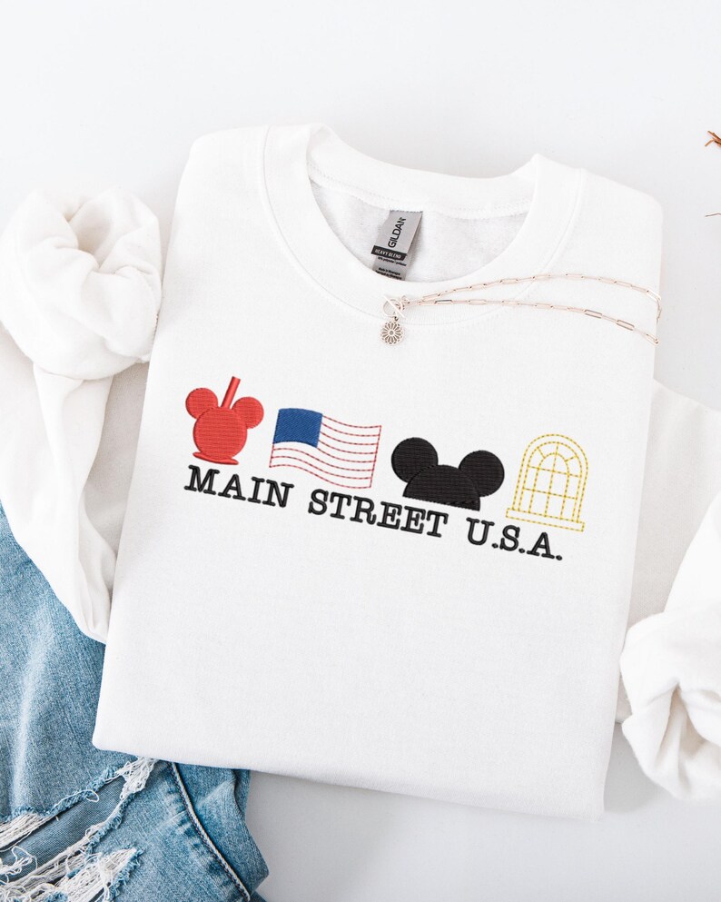 K&ouml;nnte beinhalten: Wei&szlig;es Sweatshirt mit gesticktem Main Street U.S.A. Design. Es zeigt einen roten Mickey Mouse Kopf, eine amerikanische Flagge, schwarze Mickey Mouse Ohren und ein goldenes Fenster. Eine silberne Halskette mit einem kleinen Anh&auml;nger liegt auf dem Shirt.
