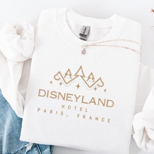 May include: A white Gildan sweatshirt with a gold Disneyland Hotel Paris, France design. A silver necklace with a small pendant rests on the folded sweatshirt. The sweatshirt is folded on top of a pair of blue jeans.