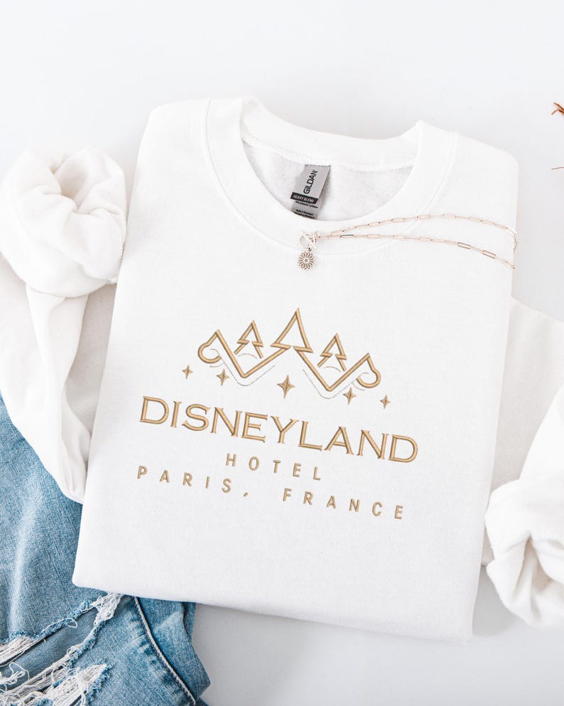 May include: A white Gildan sweatshirt with a gold Disneyland Hotel Paris, France design. A silver necklace with a small pendant rests on the folded sweatshirt. The sweatshirt is folded on top of a pair of blue jeans.