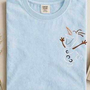 May include: Pale blue t-shirt with an embroidered Olaf design. The shirt has a Comfort Colors label. The Olaf design includes a carrot nose, stick arms, and a smiling face. The t-shirt is neatly folded.