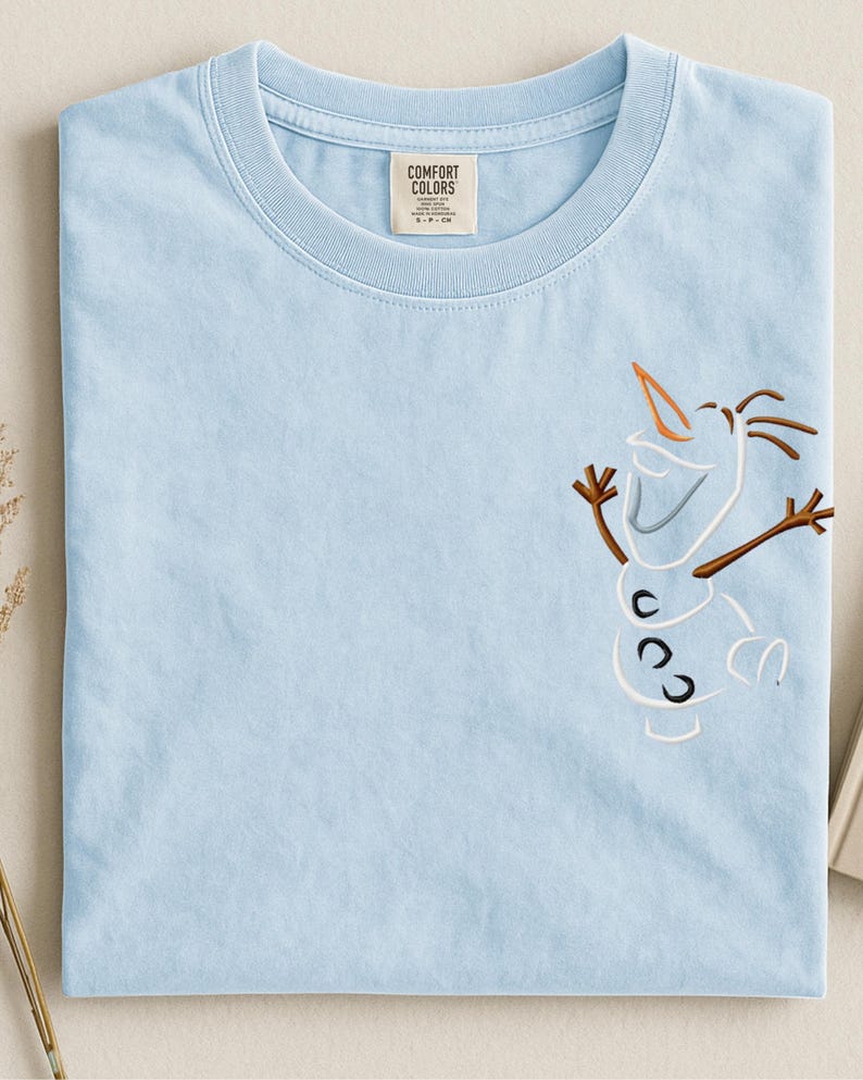 May include: Pale blue t-shirt with an embroidered Olaf design. The shirt has a Comfort Colors label. The Olaf design includes a carrot nose, stick arms, and a smiling face. The t-shirt is neatly folded.