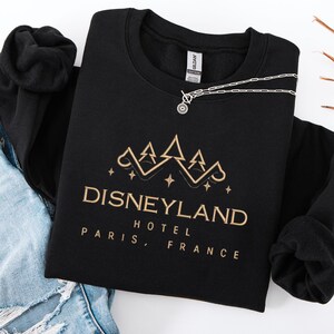 May include: Black crewneck sweatshirt with gold embroidered text and graphic. The text reads "DISNEYLAND HOTEL PARIS, FRANCE". A silver necklace with a circular pendant is draped across the sweatshirt. The sweatshirt is laid on top of a pair of blue jeans.