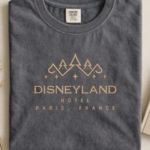 May include: Dark gray t-shirt with a gold-colored Disneyland Hotel Paris, France graphic. The design features a crown and stars. The shirt has a crew neck and a Comfort Colors tag.