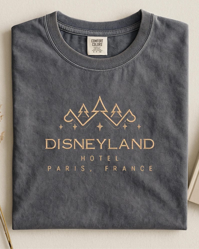 May include: Dark gray t-shirt with a gold-colored Disneyland Hotel Paris, France graphic. The design features a crown and stars. The shirt has a crew neck and a Comfort Colors tag.