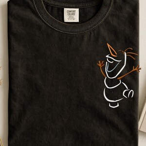 May include: Dark grey t-shirt featuring a white and brown cartoon snowman design. The shirt has a crew neck and a label that reads "Comfort Colors". The snowman has a carrot nose and stick arms.
