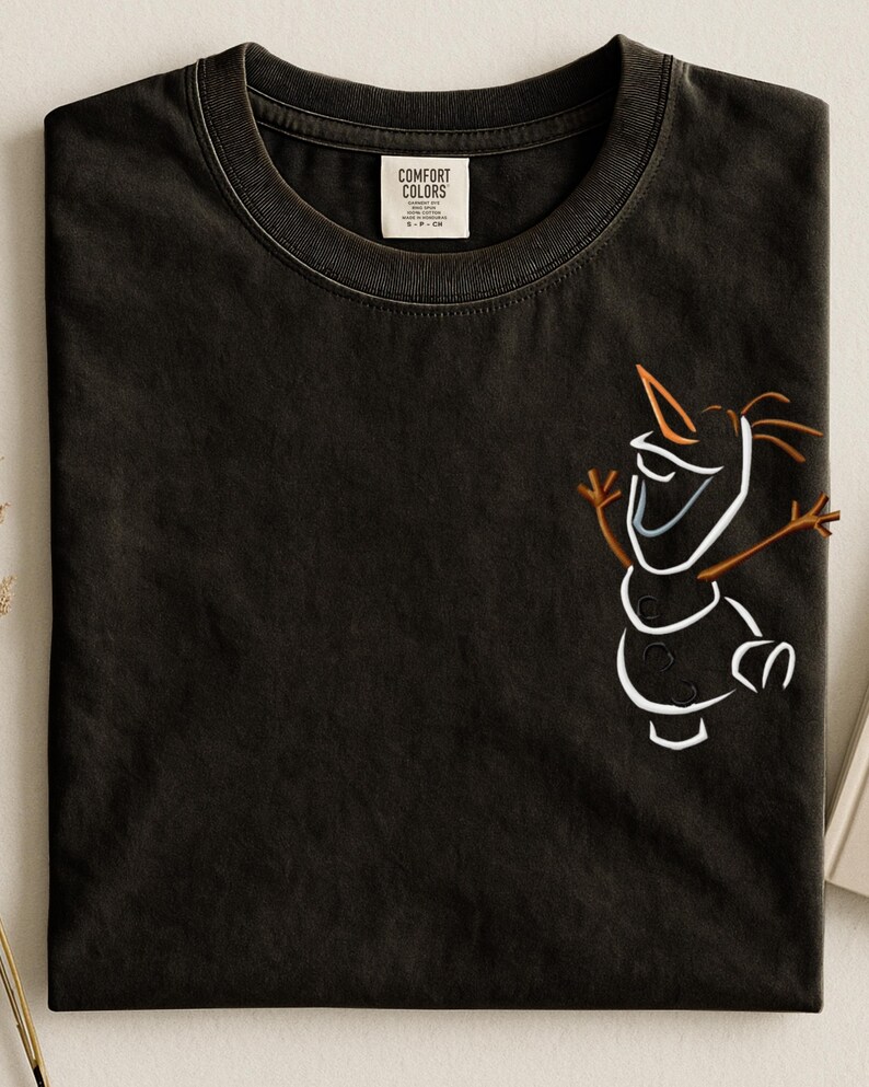 May include: Dark grey t-shirt featuring a white and brown cartoon snowman design. The shirt has a crew neck and a label that reads "Comfort Colors". The snowman has a carrot nose and stick arms.