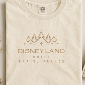 May include: Cream-colored t-shirt with a gold embroidered design featuring a crown and the text "DISNEYLAND HOTEL PARIS, FRANCE." The shirt is folded neatly, showcasing the design's details and the brand label.