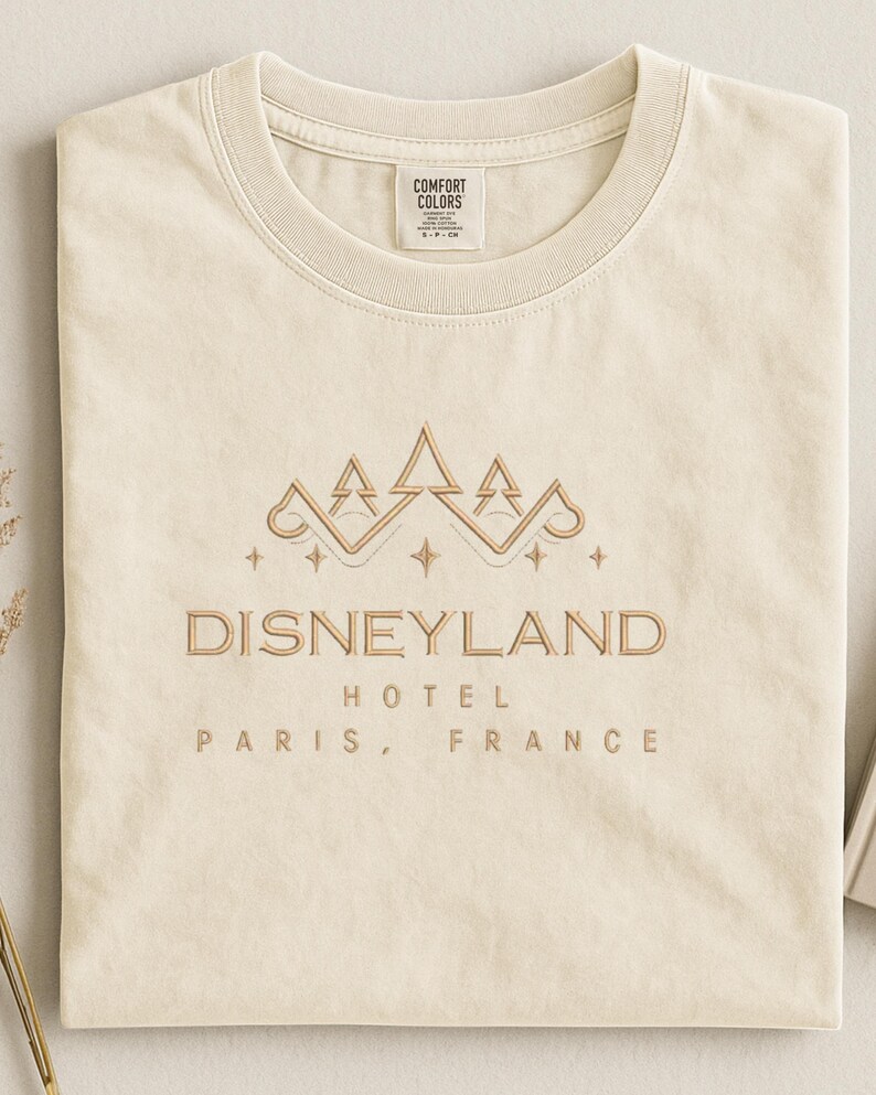 May include: Cream-colored t-shirt with a gold embroidered design featuring a crown and the text "DISNEYLAND HOTEL PARIS, FRANCE." The shirt is folded neatly, showcasing the design's details and the brand label.