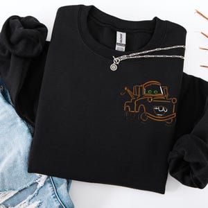 Puede incluir: Sudadera negra de cuello redondo con un dise&ntilde;o bordado de dibujos animados de gr&uacute;a en marr&oacute;n y naranja. Un collar plateado con un colgante circular descansa sobre la sudadera. La sudadera est&aacute; doblada sobre unos vaqueros azules.