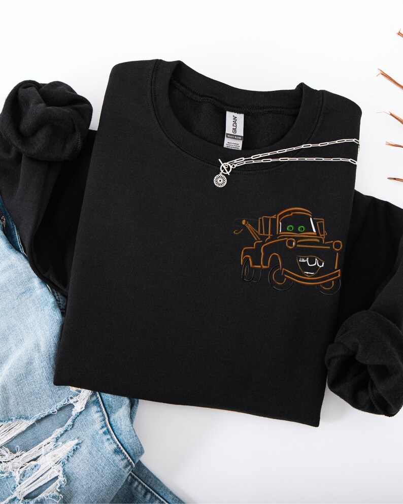 Puede incluir: Sudadera negra de cuello redondo con un dise&ntilde;o bordado de dibujos animados de gr&uacute;a en marr&oacute;n y naranja. Un collar plateado con un colgante circular descansa sobre la sudadera. La sudadera est&aacute; doblada sobre unos vaqueros azules.