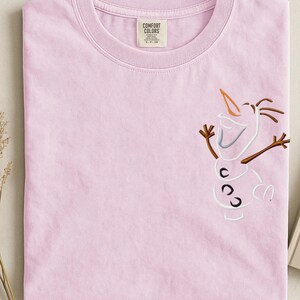 May include: A pale pink Comfort Colors t-shirt with an embroidered Olaf design. The design features a carrot nose, stick arms, and three black buttons. The shirt has a crew neck and a tag that reads "Comfort Colors."