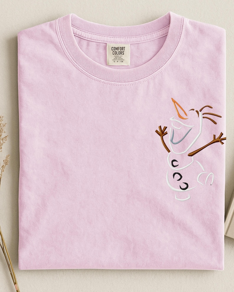 May include: A pale pink Comfort Colors t-shirt with an embroidered Olaf design. The design features a carrot nose, stick arms, and three black buttons. The shirt has a crew neck and a tag that reads "Comfort Colors."