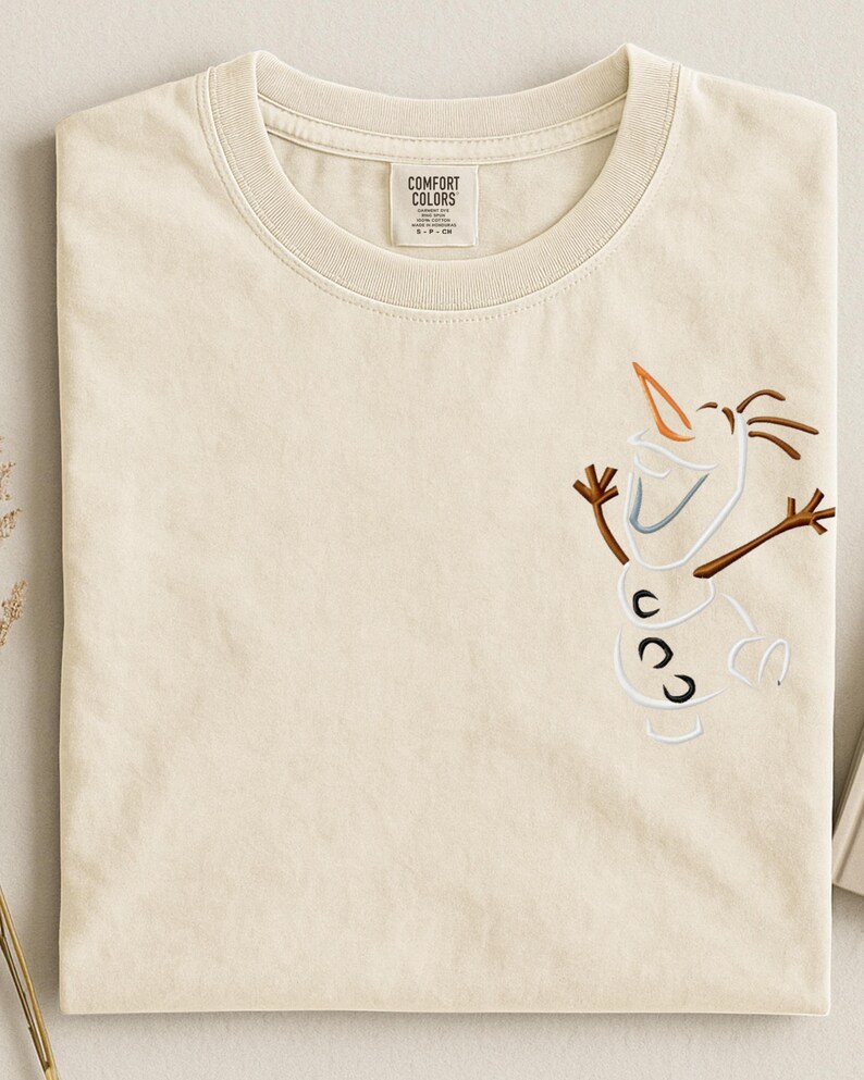 May include: Cream-coloured t-shirt with an embroidered Olaf design. The design includes a carrot nose, twig arms, and a smiling face. The shirt has a crew neck and a tag that reads "Comfort Colors."