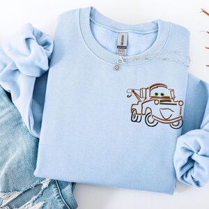 Puede incluir: Sudadera azul claro con cuello redondo con un dise&ntilde;o bordado de un cami&oacute;n de remolque de dibujos animados. La sudadera est&aacute; hecha de un material suave y se combina con un collar plateado y unos vaqueros.