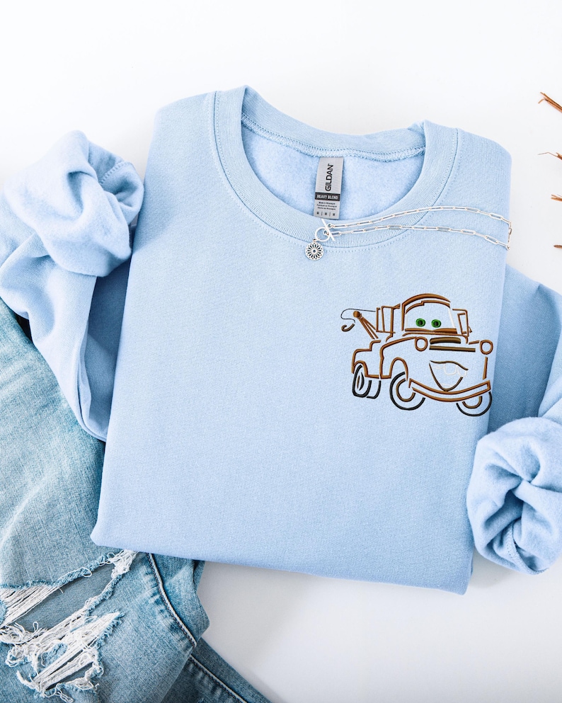Puede incluir: Sudadera azul claro con cuello redondo con un dise&ntilde;o bordado de un cami&oacute;n de remolque de dibujos animados. La sudadera est&aacute; hecha de un material suave y se combina con un collar plateado y unos vaqueros.