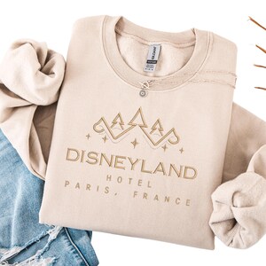 May include: Beige crewneck sweatshirt with "Disneyland Hotel Paris, France" embroidered in gold. The design includes a stylized castle and stars. A delicate necklace with a pendant is visible. The sweatshirt is folded on top of blue jeans.