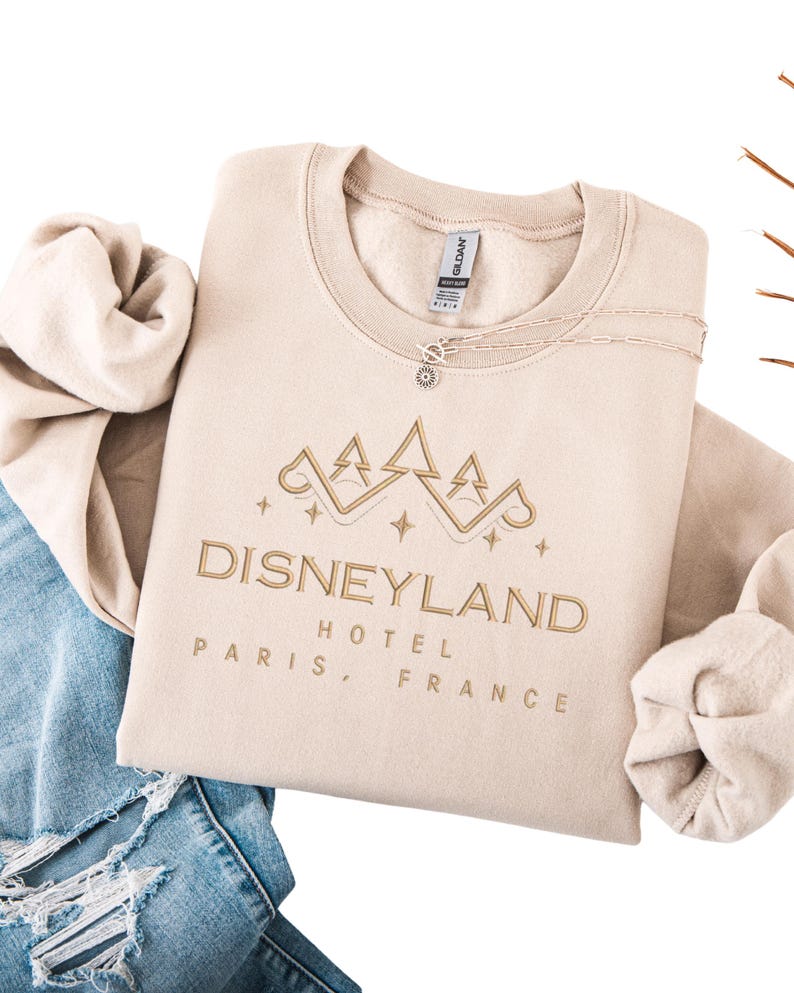 May include: Beige crewneck sweatshirt with "Disneyland Hotel Paris, France" embroidered in gold. The design includes a stylized castle and stars. A delicate necklace with a pendant is visible. The sweatshirt is folded on top of blue jeans.