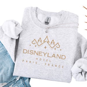 May include: A light gray sweatshirt with the text "DISNEYLAND HOTEL PARIS, FRANCE" embroidered in gold. The design includes a gold crown and stars. A silver necklace with a sun pendant rests on the sweatshirt.