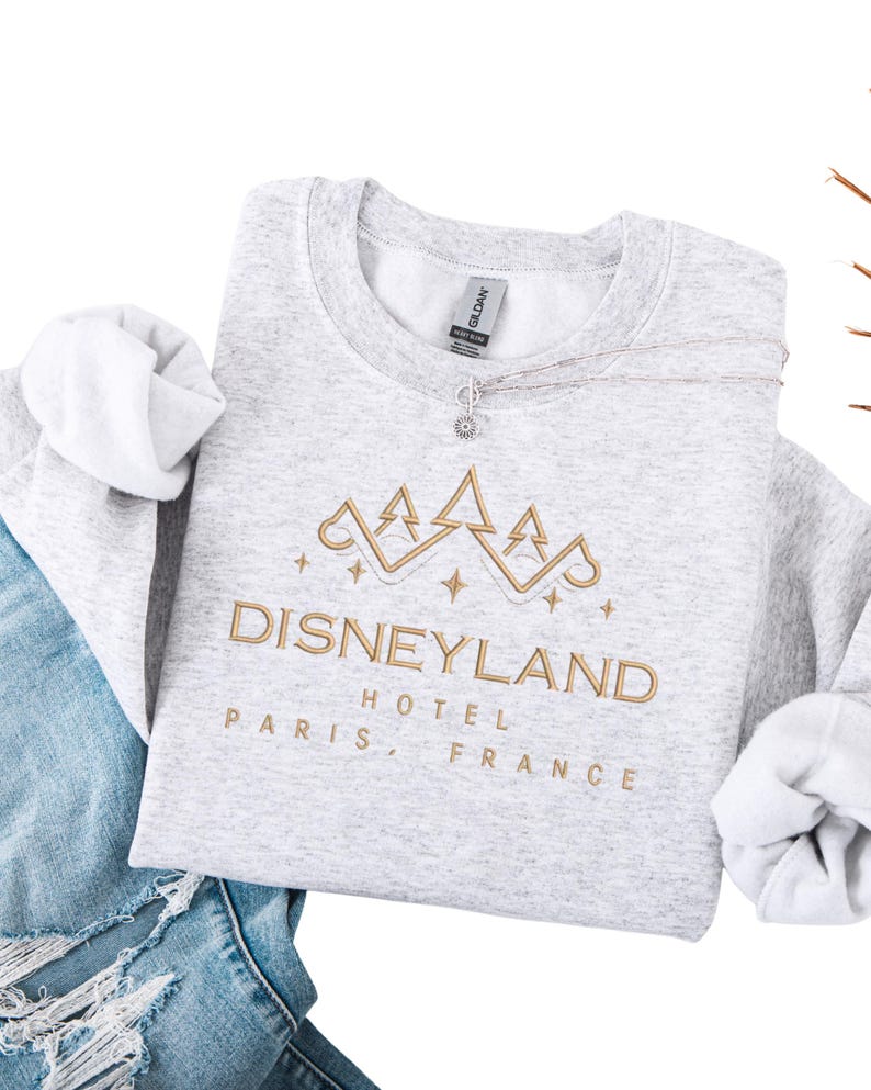 May include: A light gray sweatshirt with the text "DISNEYLAND HOTEL PARIS, FRANCE" embroidered in gold. The design includes a gold crown and stars. A silver necklace with a sun pendant rests on the sweatshirt.