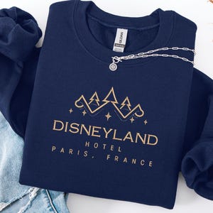 May include: A navy blue sweatshirt with gold text that reads "Disneyland Hotel Paris, France." A silver necklace with a sun-shaped pendant rests on the sweatshirt. The sweatshirt is folded on top of a pair of blue jeans.