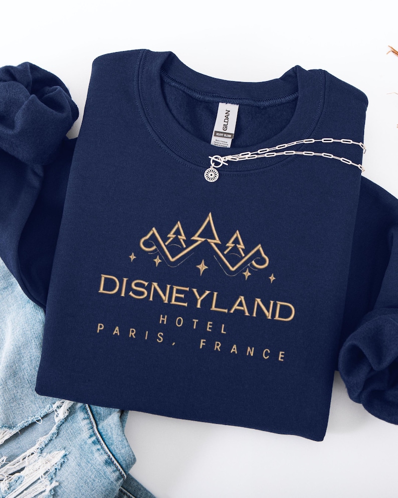 May include: A navy blue sweatshirt with gold text that reads "Disneyland Hotel Paris, France." A silver necklace with a sun-shaped pendant rests on the sweatshirt. The sweatshirt is folded on top of a pair of blue jeans.