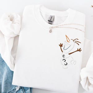 May include: White crew neck sweatshirt with an embroidered Olaf design. The design features brown twig arms, a carrot nose, and a smiling face. A silver necklace with a small pendant rests on the sweatshirt. The sweatshirt is folded on a white surface.