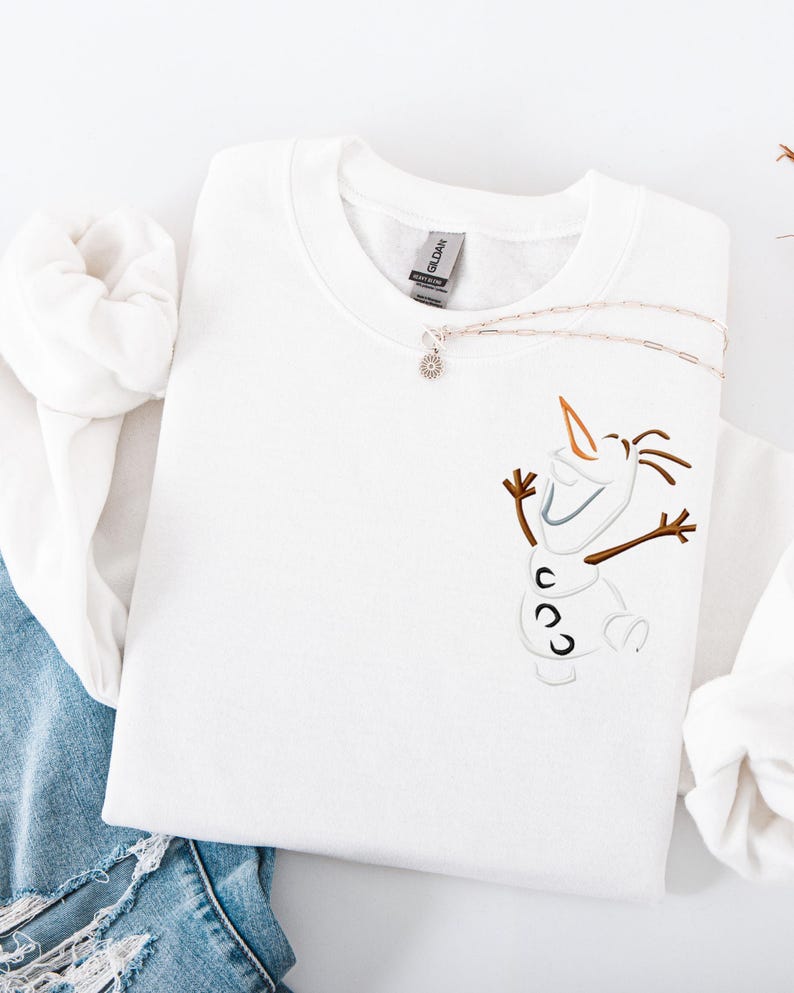 May include: White crew neck sweatshirt with an embroidered Olaf design. The design features brown twig arms, a carrot nose, and a smiling face. A silver necklace with a small pendant rests on the sweatshirt. The sweatshirt is folded on a white surface.