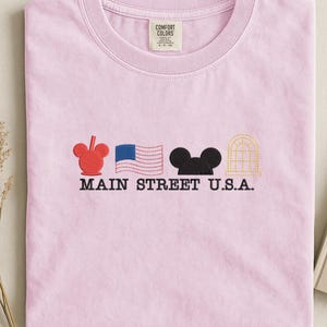 K&ouml;nnte beinhalten: Ein hellrosa T-Shirt mit gestickten Designs und dem Text "MAIN STREET U.S.A.". Es zeigt einen roten Mickey Mouse-Kopf, eine blau-wei&szlig;e amerikanische Flagge, eine schwarze Mickey Mouse-Silhouette und ein gelbes Bogenfenster.