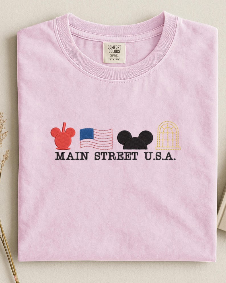 K&ouml;nnte beinhalten: Ein hellrosa T-Shirt mit gestickten Designs und dem Text "MAIN STREET U.S.A.". Es zeigt einen roten Mickey Mouse-Kopf, eine blau-wei&szlig;e amerikanische Flagge, eine schwarze Mickey Mouse-Silhouette und ein gelbes Bogenfenster.
