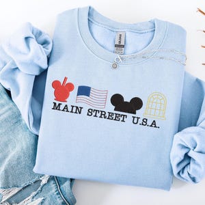 K&ouml;nnte beinhalten: Hellblaues Sweatshirt mit gesticktem Main Street U.S.A. Design. Es zeigt einen roten Mickey Mouse-Kopf, eine amerikanische Flagge, einen schwarzen Mickey Mouse-Kopf und ein gelbes Fenster. Eine silberne Halskette liegt auf dem Sweatshirt.