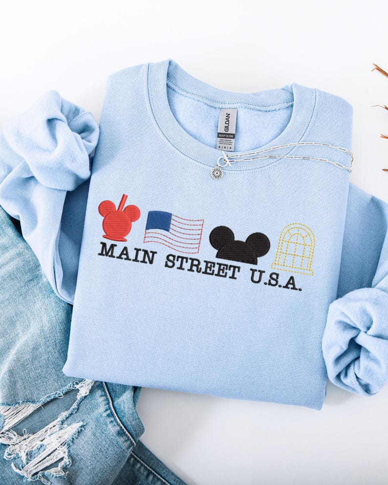K&ouml;nnte beinhalten: Hellblaues Sweatshirt mit gesticktem Main Street U.S.A. Design. Es zeigt einen roten Mickey Mouse-Kopf, eine amerikanische Flagge, einen schwarzen Mickey Mouse-Kopf und ein gelbes Fenster. Eine silberne Halskette liegt auf dem Sweatshirt.