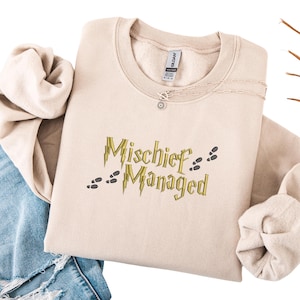 May include: A beige crew neck sweatshirt featuring the embroidered phrase "Mischief Managed" in gold, with lightning bolts and footprints. A silver necklace with a pendant is visible. The sweatshirt is folded on blue denim jeans.