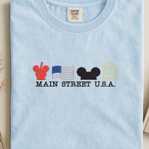 K&ouml;nnte beinhalten: Hellblaues T-Shirt mit Stickereien: roter Mickey Mouse Kopf, US-Flagge, schwarzer Mickey Mouse Kopf und ein goldenes Geb&auml;ude. Der Text "MAIN STREET U.S.A." ist darunter gestickt. Mit Comfort Colors Etikett.
