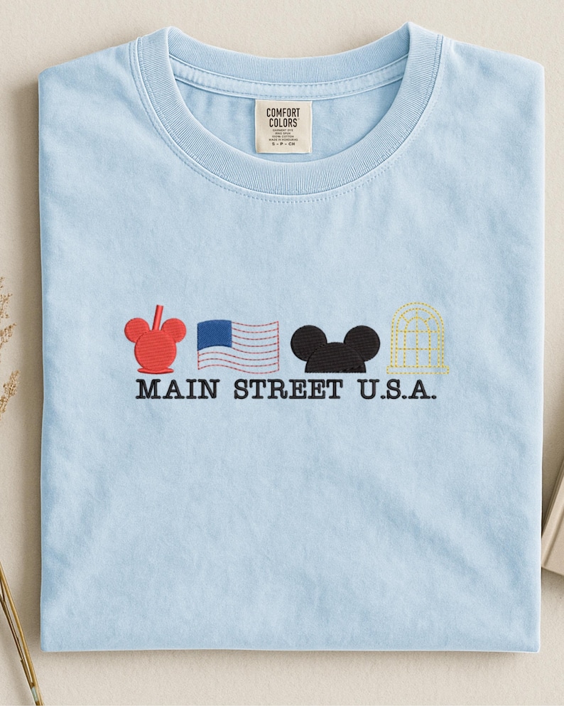 K&ouml;nnte beinhalten: Hellblaues T-Shirt mit Stickereien: roter Mickey Mouse Kopf, US-Flagge, schwarzer Mickey Mouse Kopf und ein goldenes Geb&auml;ude. Der Text "MAIN STREET U.S.A." ist darunter gestickt. Mit Comfort Colors Etikett.