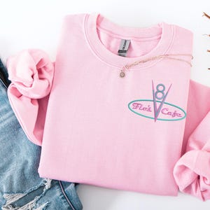 Puede incluir: Sudadera rosa claro de cuello redondo con el texto bordado "Flo's Cafe" en un estilo retro de restaurante. El dise&ntilde;o incluye un &oacute;valo turquesa, l&iacute;neas rosas y el n&uacute;mero "8". Un delicado collar con un peque&ntilde;o colgante descansa sobre la sudadera.