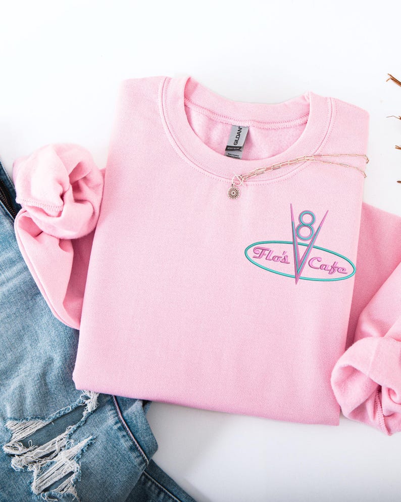 Puede incluir: Sudadera rosa claro de cuello redondo con el texto bordado "Flo's Cafe" en un estilo retro de restaurante. El dise&ntilde;o incluye un &oacute;valo turquesa, l&iacute;neas rosas y el n&uacute;mero "8". Un delicado collar con un peque&ntilde;o colgante descansa sobre la sudadera.