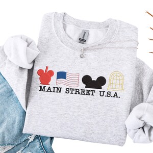 K&ouml;nnte beinhalten: Ein hellgraues Sweatshirt mit gestickten Designs und dem Text "MAIN STREET U.S.A.". Es zeigt einen roten Mickey Mouse-Kopf, eine amerikanische Flagge, einen schwarzen Mickey Mouse-Kopf und ein goldfarbenes Bogenfenster. Das Sweatshirt wird mit einer blauen Jeans kombiniert.