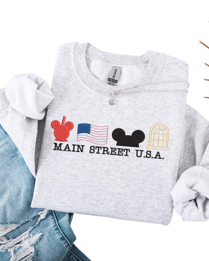 K&ouml;nnte beinhalten: Ein hellgraues Sweatshirt mit gestickten Designs und dem Text "MAIN STREET U.S.A.". Es zeigt einen roten Mickey Mouse-Kopf, eine amerikanische Flagge, einen schwarzen Mickey Mouse-Kopf und ein goldfarbenes Bogenfenster. Das Sweatshirt wird mit einer blauen Jeans kombiniert.