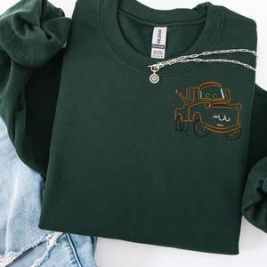 Puede incluir: Sudadera verde oscuro con un dise&ntilde;o bordado de un cami&oacute;n de remolque de dibujos animados. La sudadera tiene cuello redondo y mangas largas. Un collar plateado con un colgante de sol est&aacute; drapeado en la parte delantera. La sudadera se combina con unos vaqueros azules.